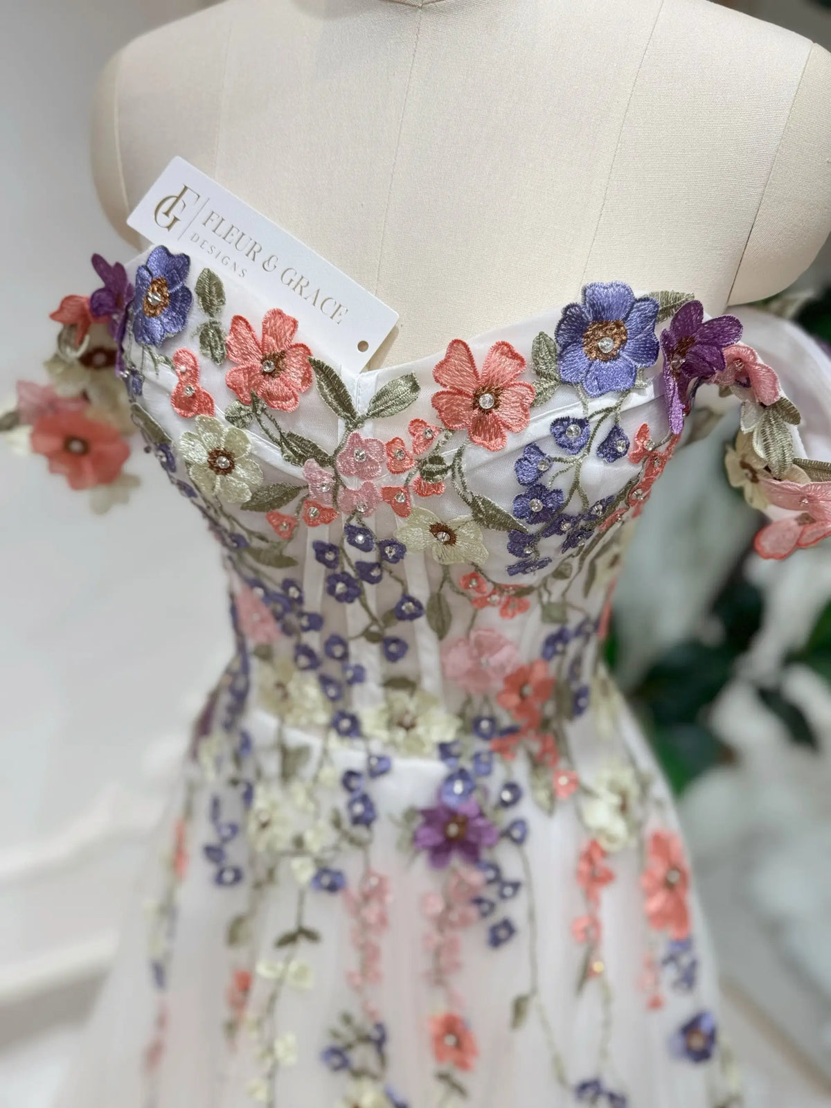 Off-shoulder wedding dress with colorful 3D floral appliqué, beaded accents, and a structured corset bodice.