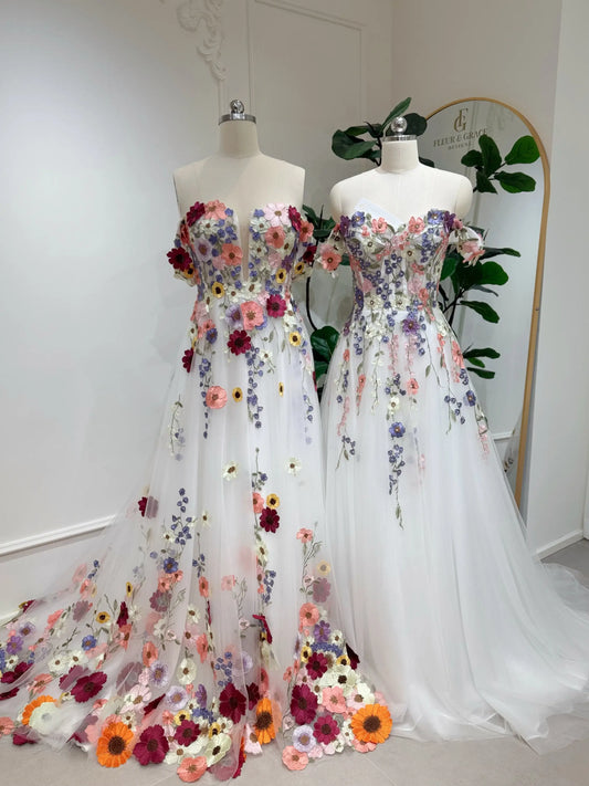 Two off-shoulder A-line wedding dresses in white tulle with colorful 3D floral appliqué, beading, and sequin details.