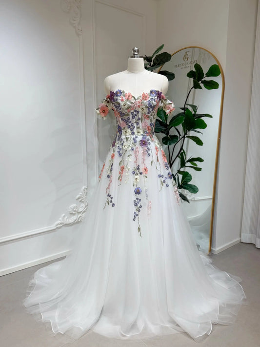 Off-shoulder A-line wedding dress with colorful 3D floral appliqué, beaded details, and a flowing white tulle skirt.