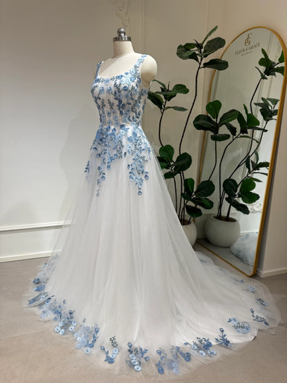 White tulle wedding gown with dusty blue floral embroidery, square neckline, and a flowing chapel train on a mannequin.
