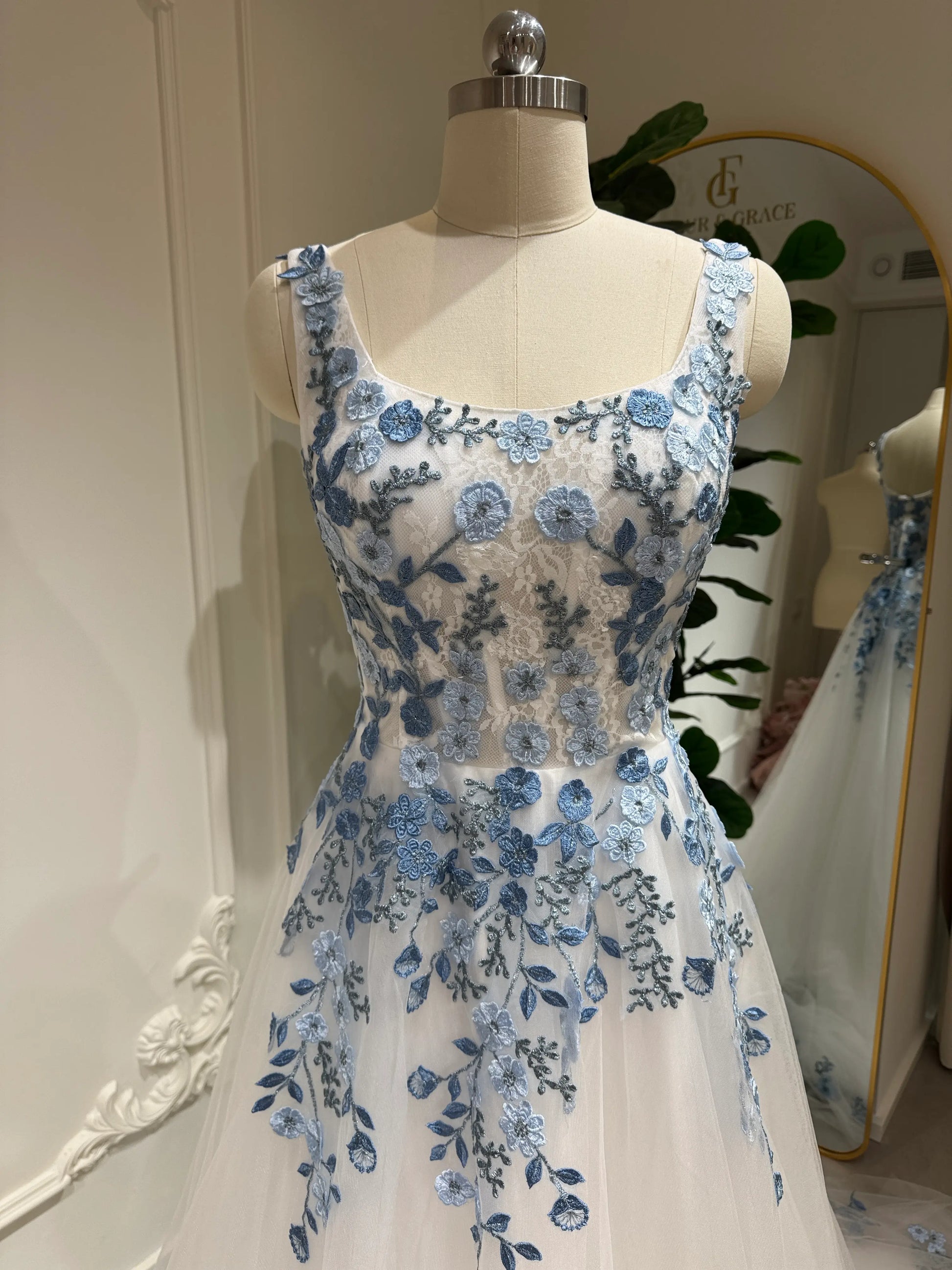 White tulle wedding gown with dusty blue floral embroidery, square neckline, and sleeveless bodice on a mannequin.