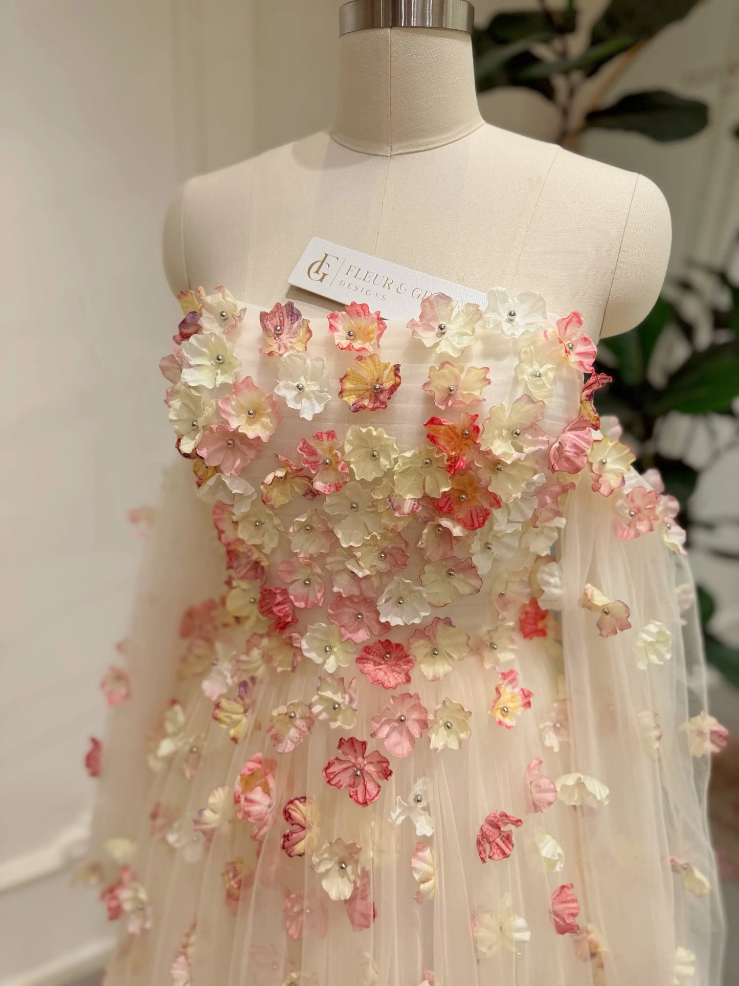 Pink strapless wedding gown featuring 3D floral appliqués, a structured bodice, and sheer removable sleeves on a mannequin.