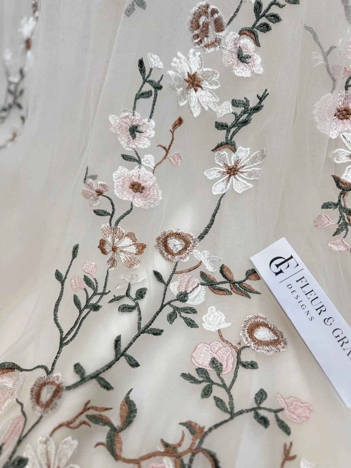 Champagne tulle fabric featuring delicate multicolored floral embroidery and a Fleur&Grace Designs brand tag.