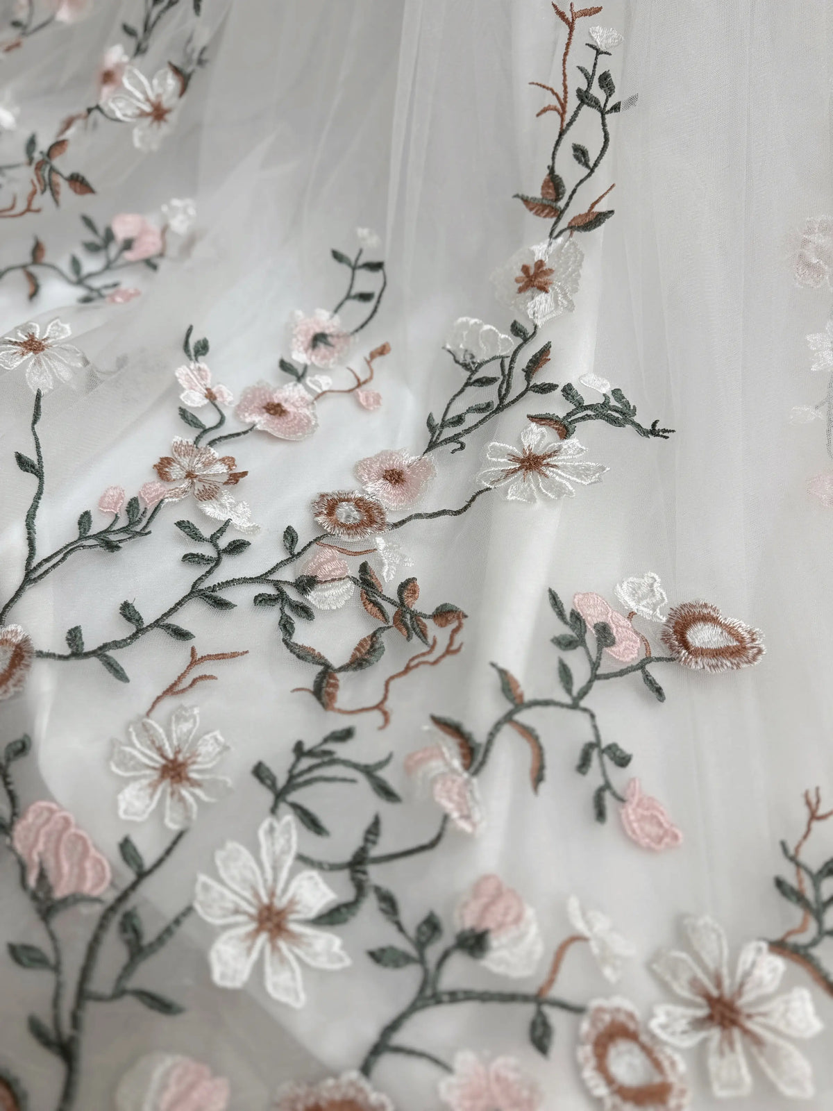 White tulle wedding gown fabric featuring delicate pink and green meadow-inspired floral embroidery.