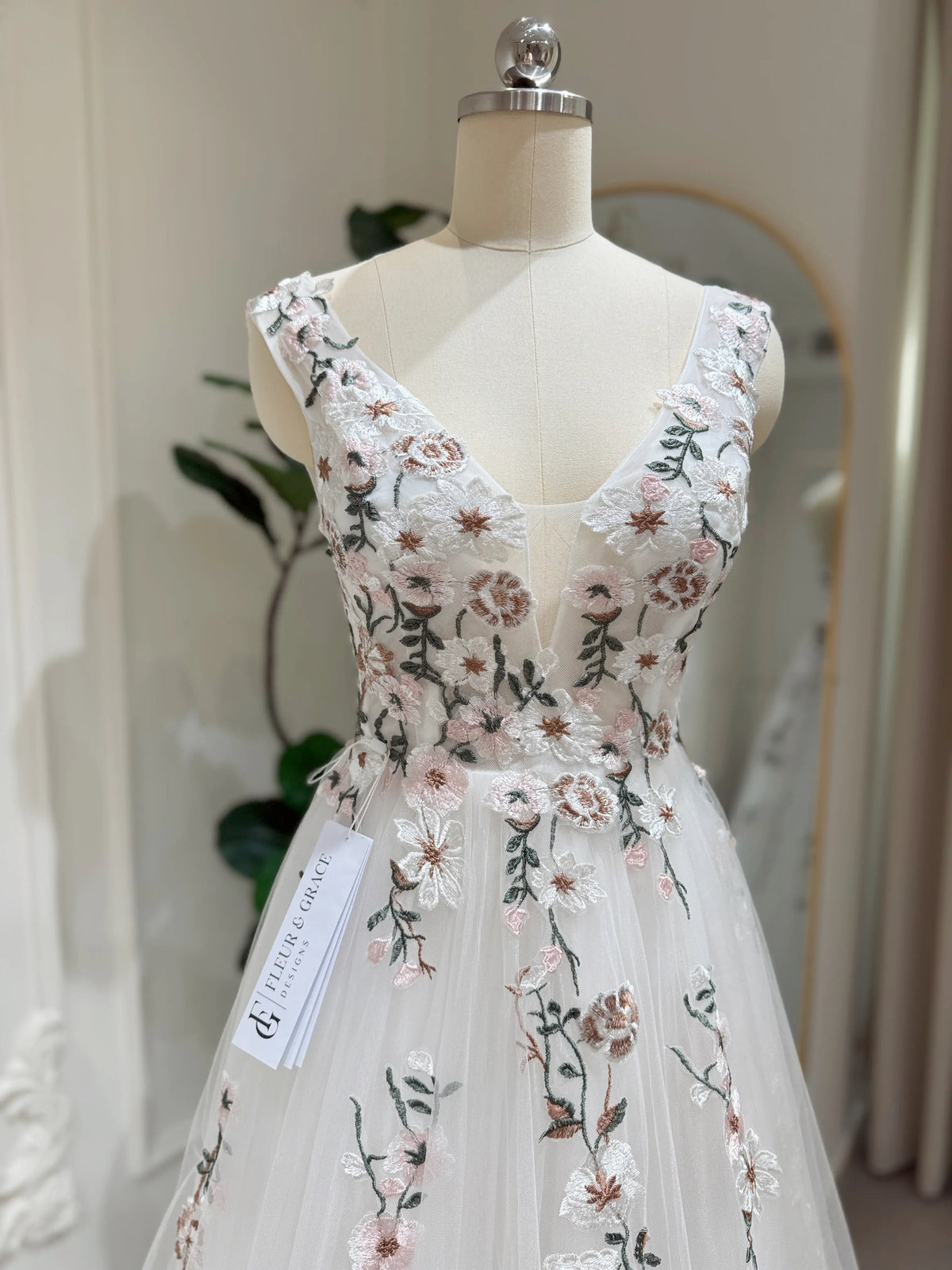 White tulle wedding gown with V-neckline and pink and green floral embroidery on a dress form.