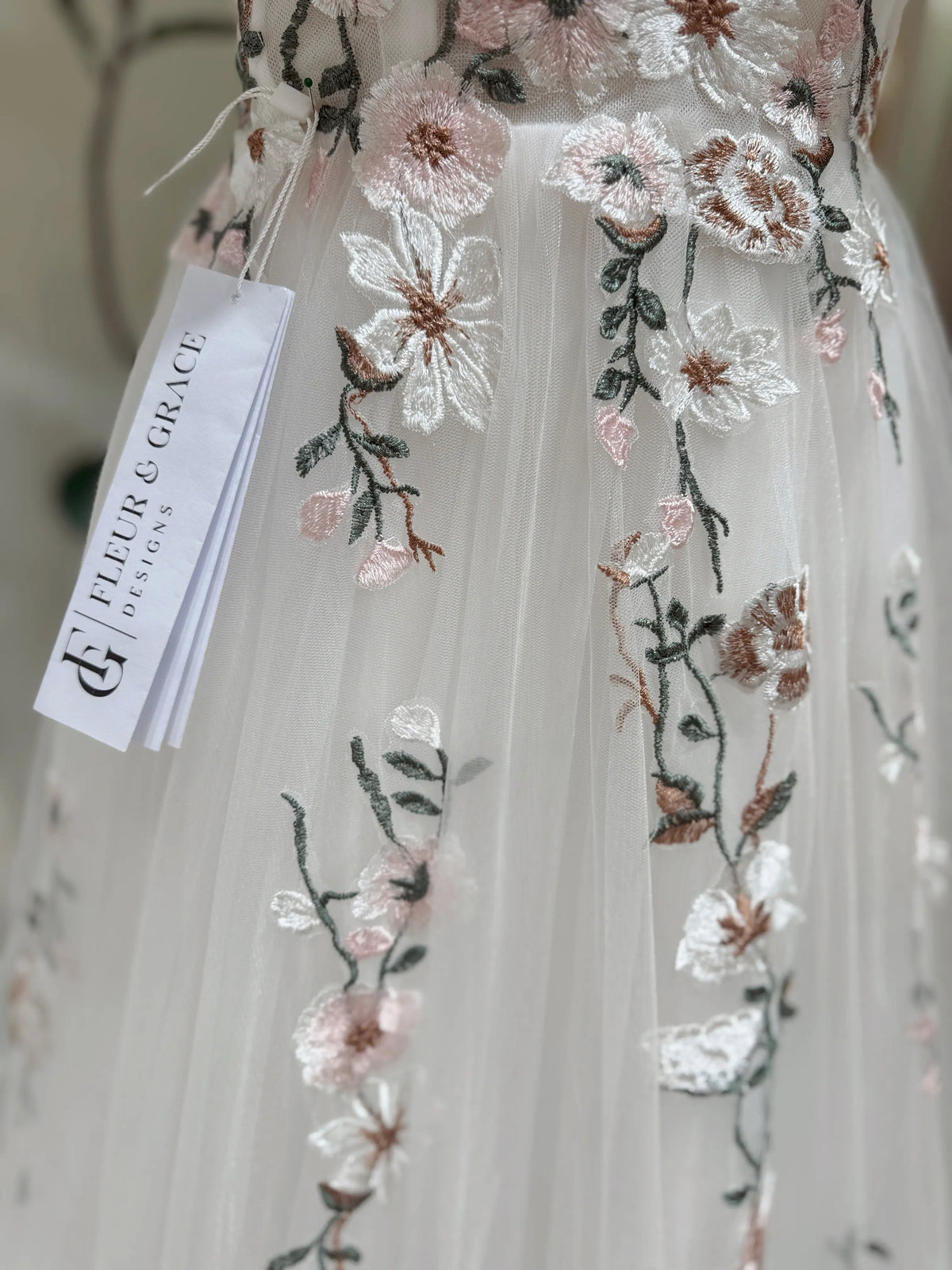 White tulle wedding gown featuring delicate pink and green floral embroidery with a Fleur & Grace brand tag.