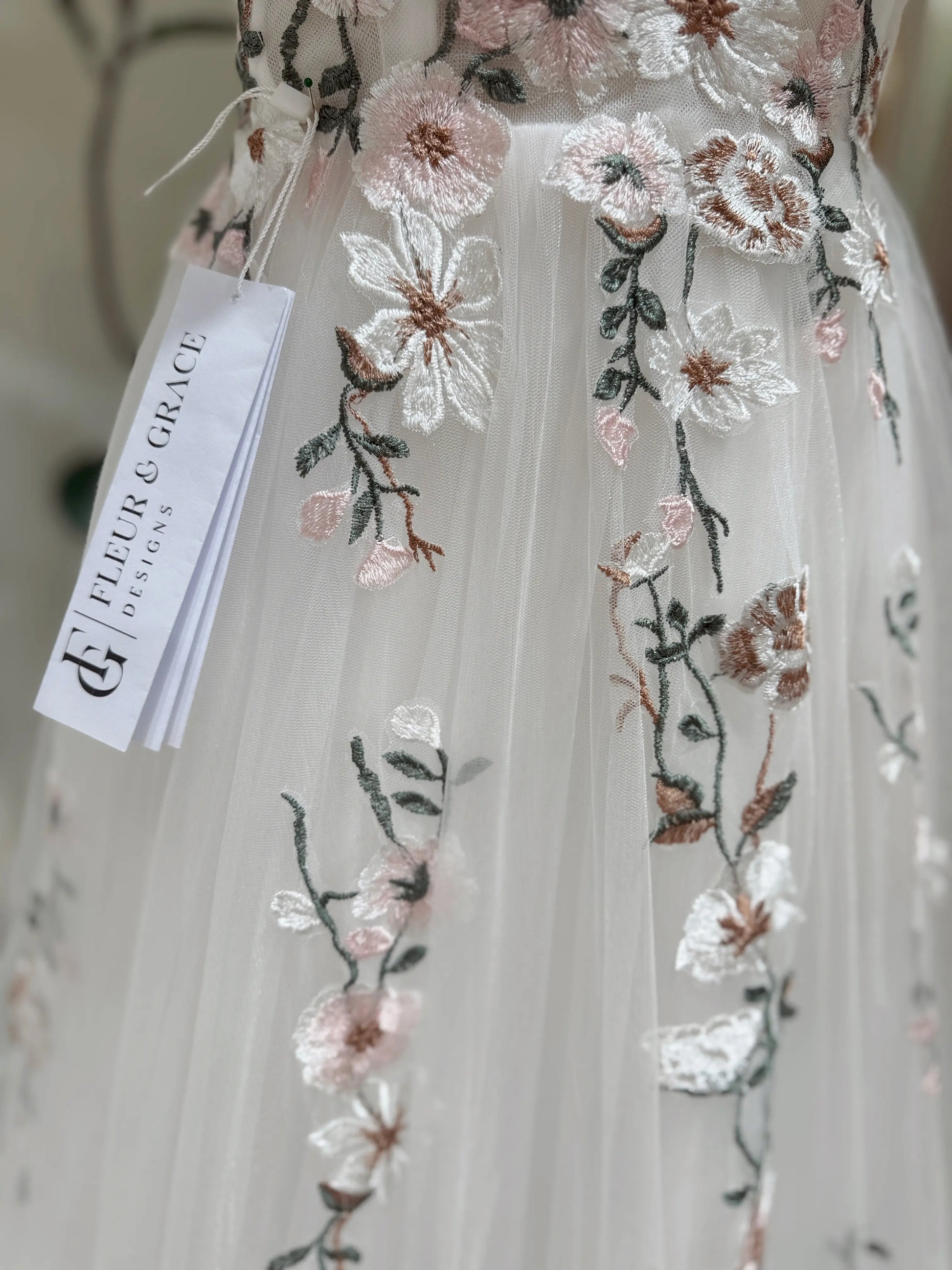 White tulle wedding gown featuring delicate pink and green floral embroidery with a Fleur & Grace brand tag.