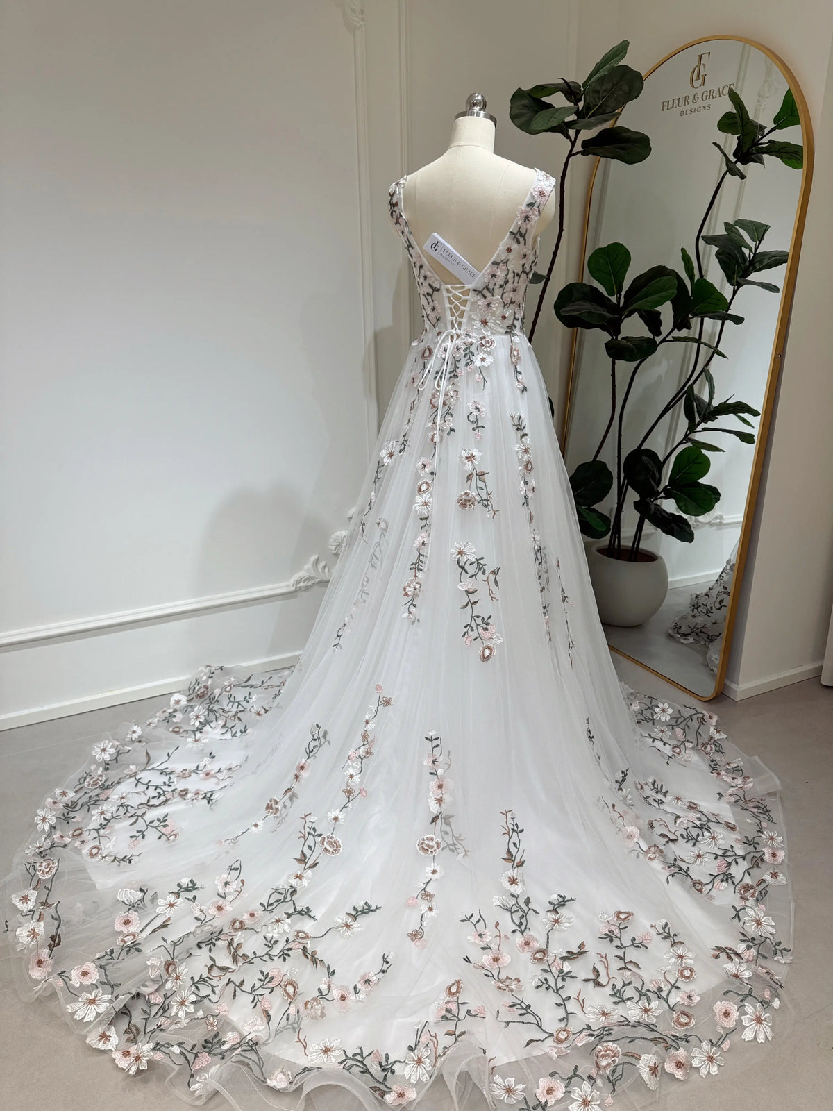 White tulle wedding gown with pink and green floral embroidery, lace-up back, and chapel train on a mannequin.