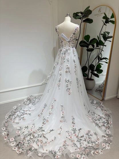 White tulle wedding gown with pink and green floral embroidery, lace-up back, and chapel train on a mannequin.