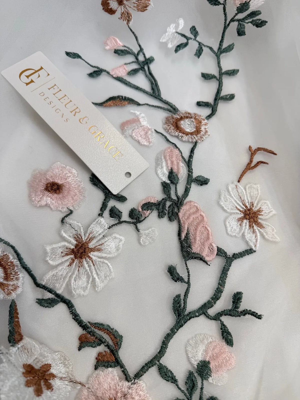 Close-up of white tulle wedding dress fabric featuring delicate 3D floral embroidery in pink, white, and sage green.
