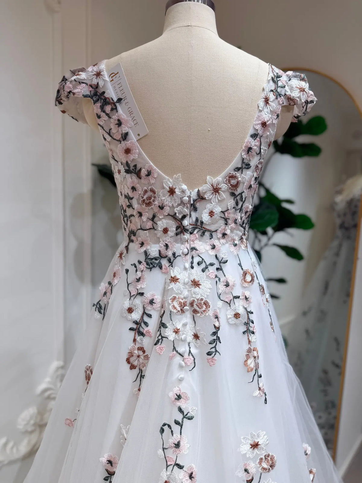 White A-line wedding dress with cap sleeves, featuring pink and green 3D floral embroidery and a button-back bodice.