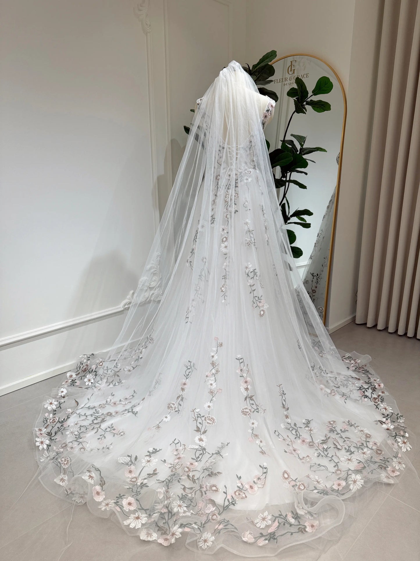 Bride mannequin wearing the Kate Gown by Fleur&Grace Designs – white tulle wedding dress with meadow floral embroidery, V‑neckline, lace‑up back, pockets, and chapel‑length train.