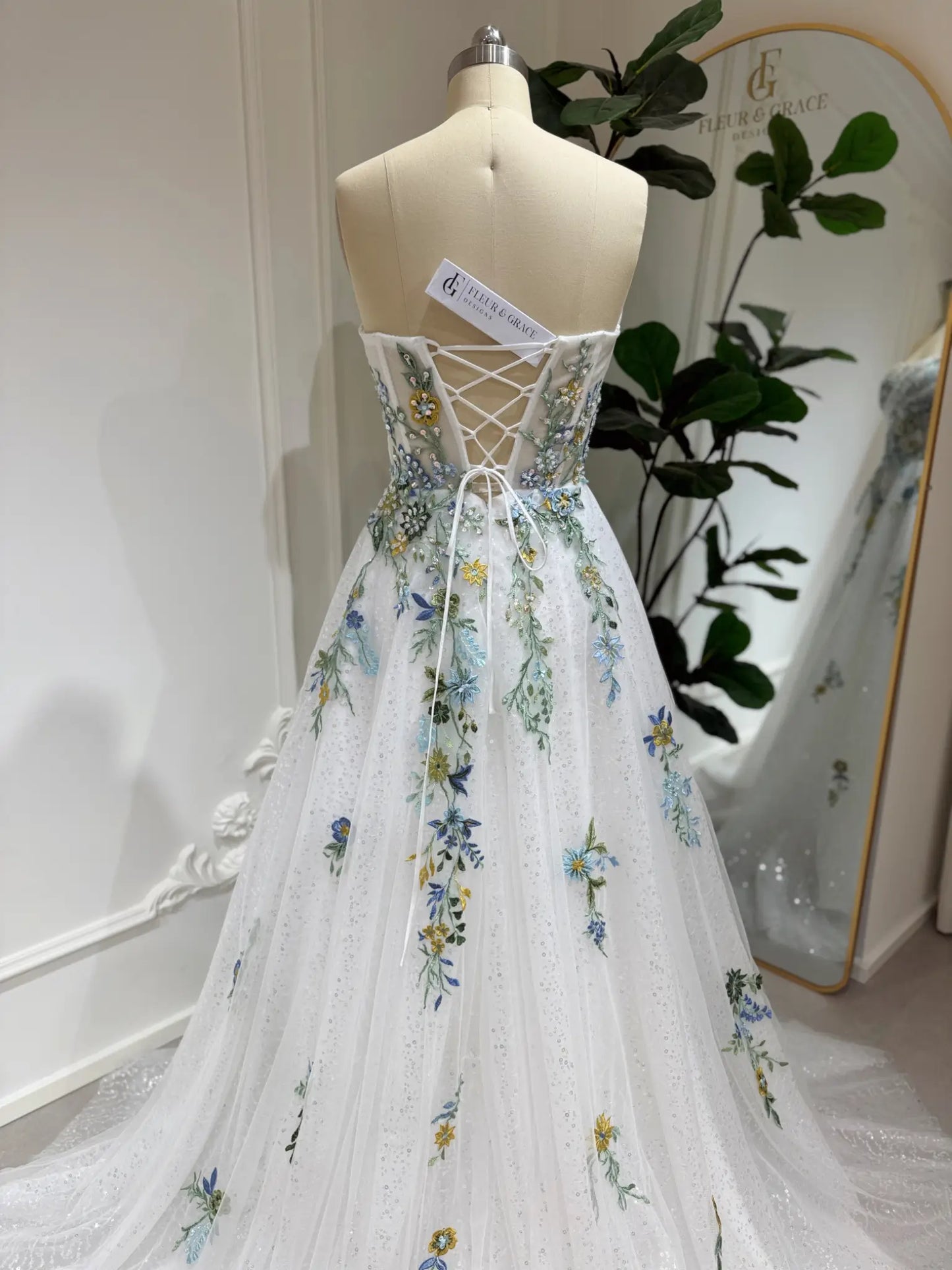 Whimsical white wedding gown with colorful floral embroidery, sparkly tulle, and a lace-up corset back on a mannequin.