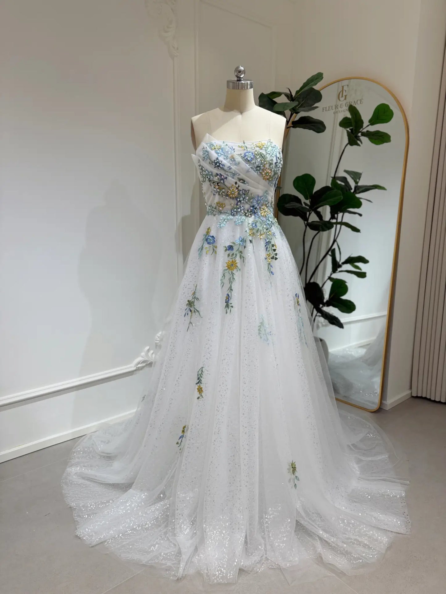 Whimsical white wedding gown with colorful floral embroidery, sparkly tulle, and a flowing skirt on a mannequin.