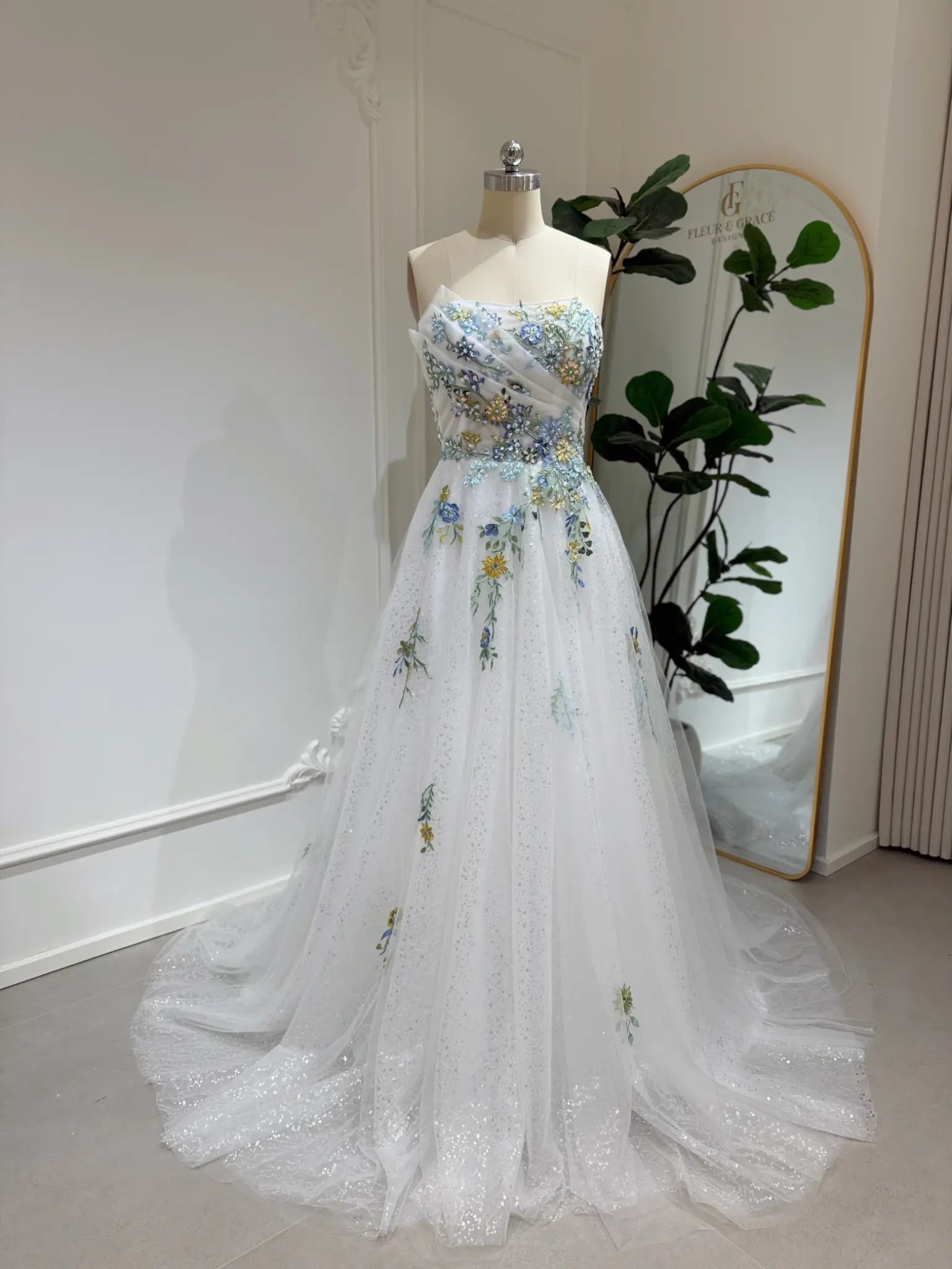 Whimsical white wedding gown with colorful floral embroidery, sparkly tulle, and a flowing skirt on a mannequin.