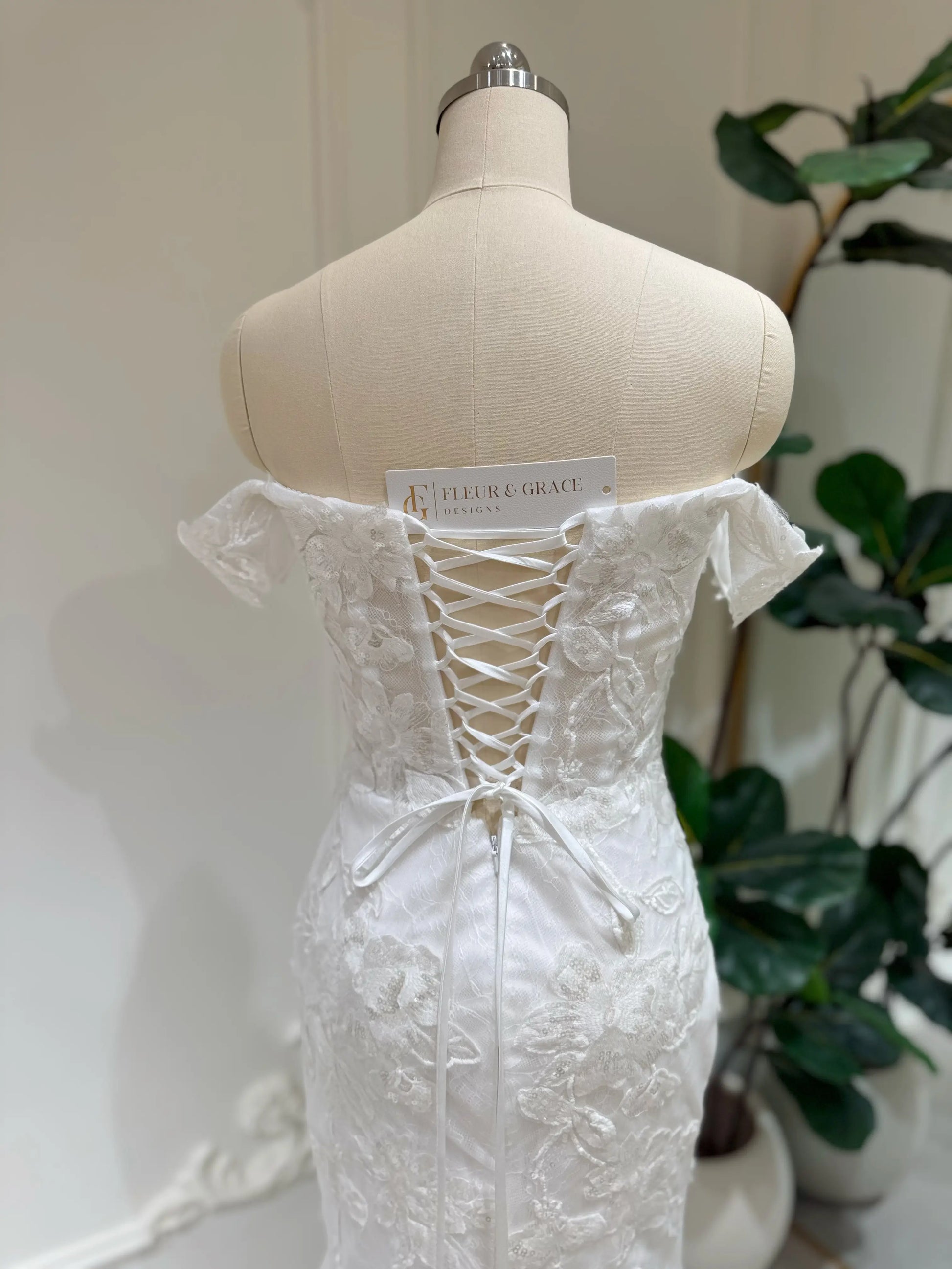 Bridal mannequin wearing the Lena Gown FG030 by Fleur&Grace Designs—a mermaid lace wedding dress with off-the-shoulder sleeves, sheer boned bodice, corset back, basque waistline, asymmetric hemline, and chapel-length train.