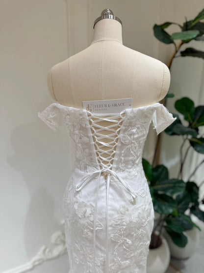 Bridal mannequin wearing the Lena Gown FG030 by Fleur&Grace Designs—a mermaid lace wedding dress with off-the-shoulder sleeves, sheer boned bodice, corset back, basque waistline, asymmetric hemline, and chapel-length train.