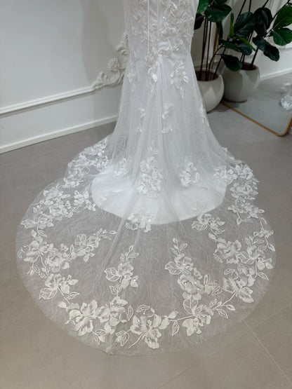 Bridal mannequin wearing the Lena Gown FG030 by Fleur&Grace Designs—a mermaid lace wedding dress with off-the-shoulder sleeves, sheer boned bodice, corset back, basque waistline, asymmetric hemline, and chapel-length train.