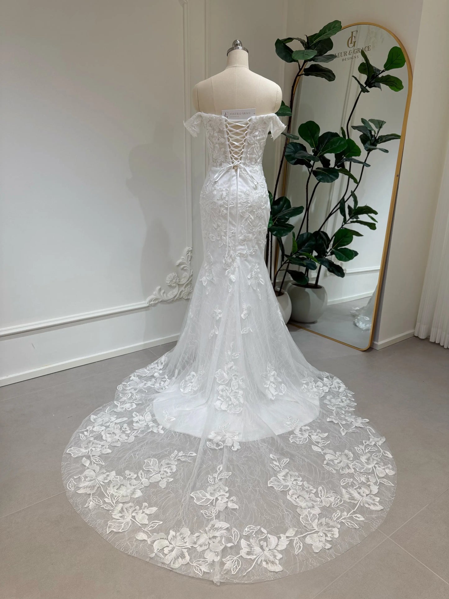 Bridal mannequin wearing the Lena Gown FG030 by Fleur&Grace Designs—a mermaid lace wedding dress with off-the-shoulder sleeves, sheer boned bodice, corset back, basque waistline, asymmetric hemline, and chapel-length train.