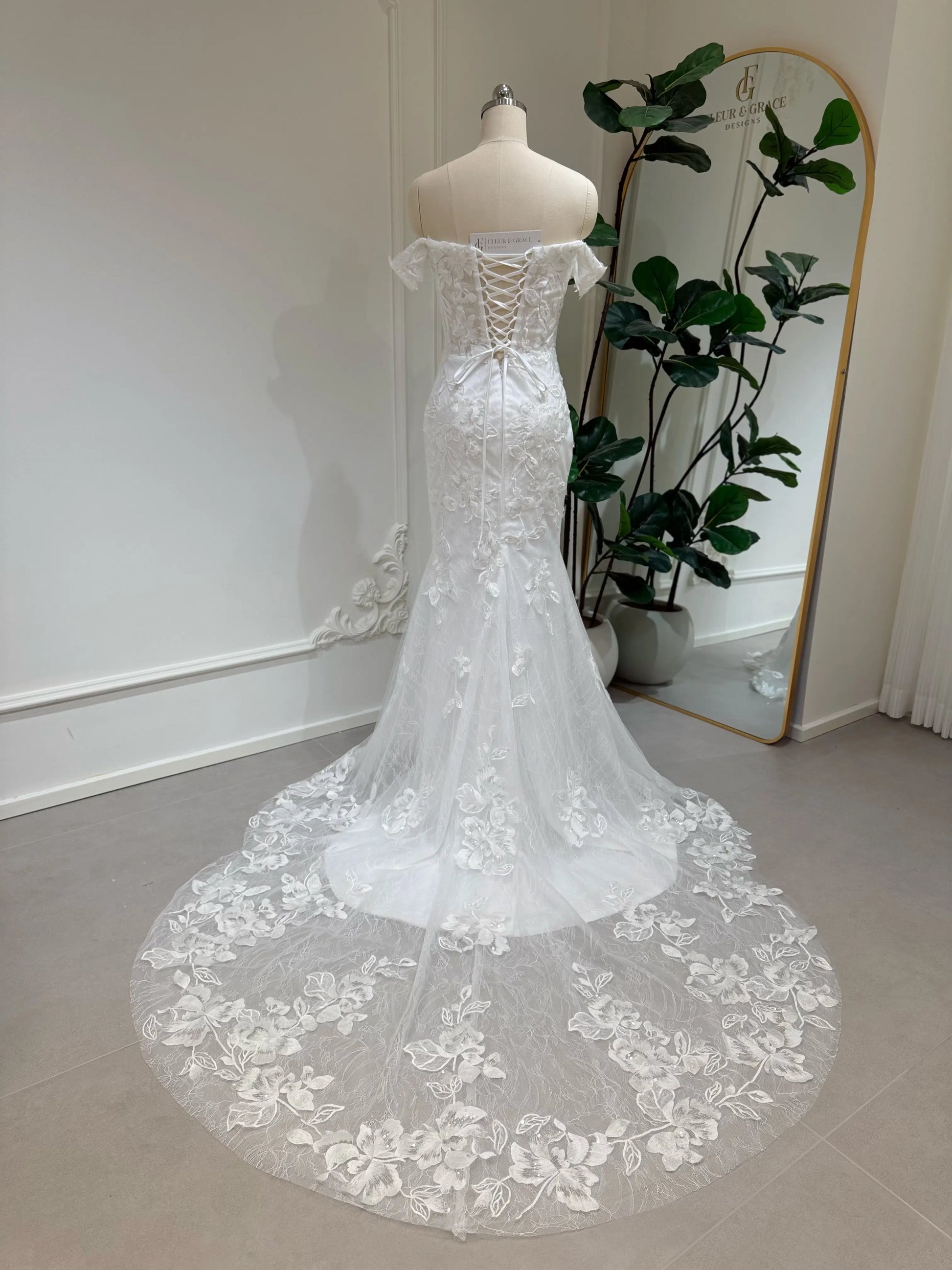 Bridal mannequin wearing the Lena Gown FG030 by Fleur&Grace Designs—a mermaid lace wedding dress with off-the-shoulder sleeves, sheer boned bodice, corset back, basque waistline, asymmetric hemline, and chapel-length train.
