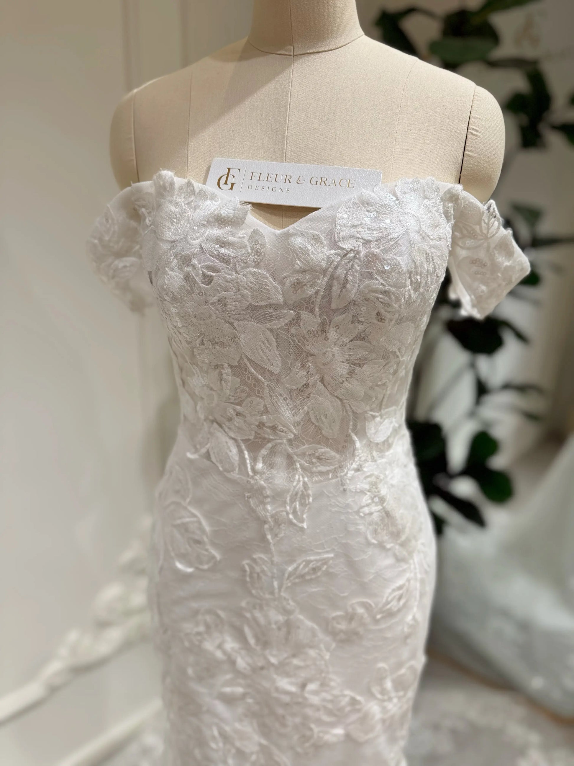Bridal mannequin wearing the Lena Gown FG030 by Fleur&Grace Designs—a mermaid lace wedding dress with off-the-shoulder sleeves, sheer boned bodice, corset back, basque waistline, asymmetric hemline, and chapel-length train.