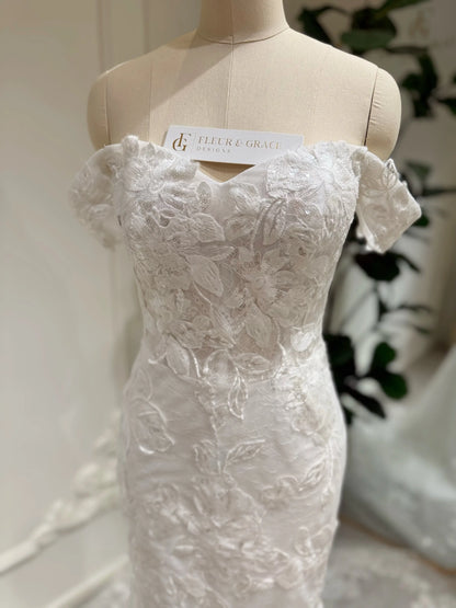 Bridal mannequin wearing the Lena Gown FG030 by Fleur&Grace Designs—a mermaid lace wedding dress with off-the-shoulder sleeves, sheer boned bodice, corset back, basque waistline, asymmetric hemline, and chapel-length train.