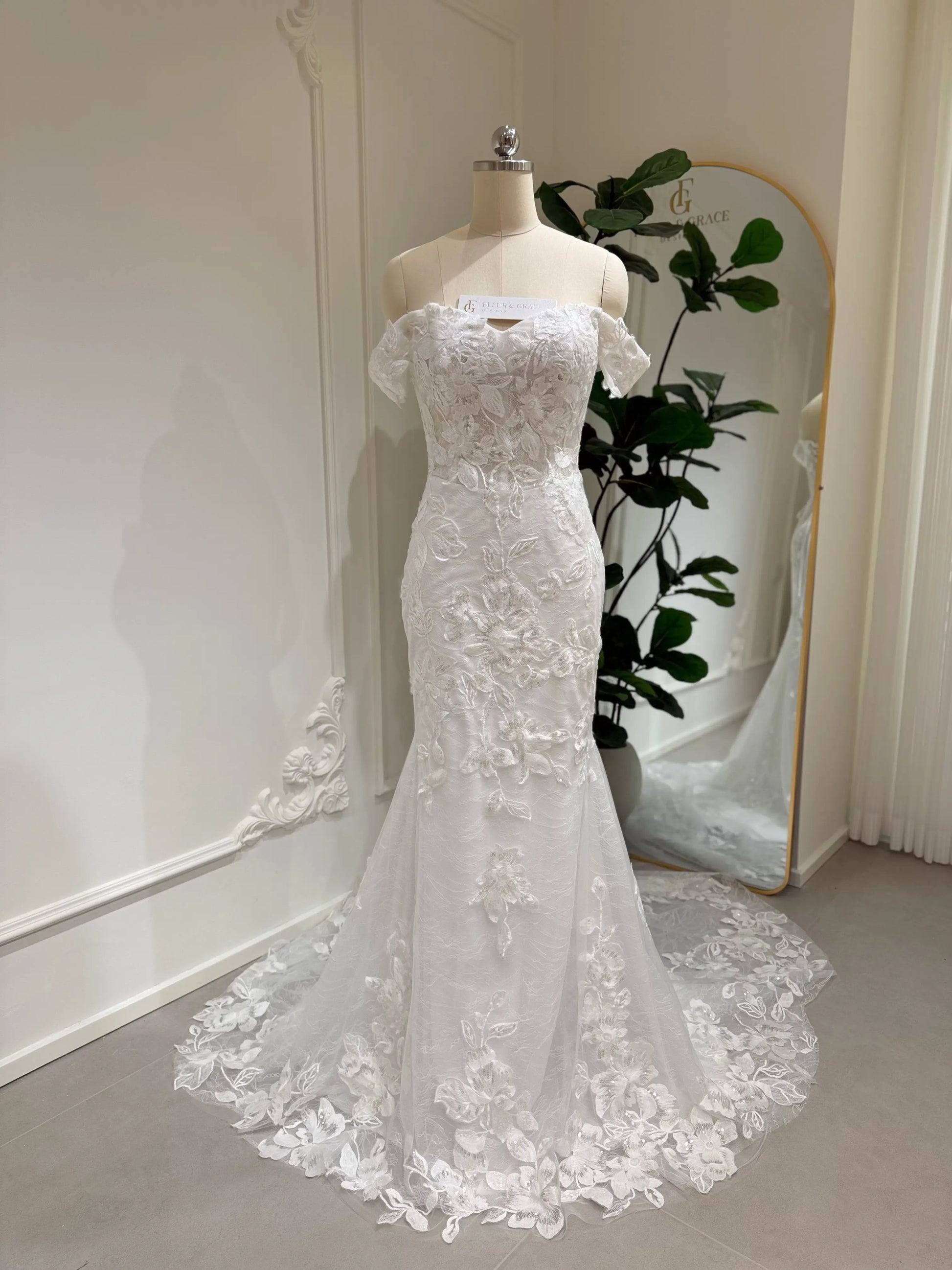 Bridal mannequin wearing the Lena Gown FG030 by Fleur&Grace Designs—a mermaid lace wedding dress with off-the-shoulder sleeves, sheer boned bodice, corset back, basque waistline, asymmetric hemline, and chapel-length train.