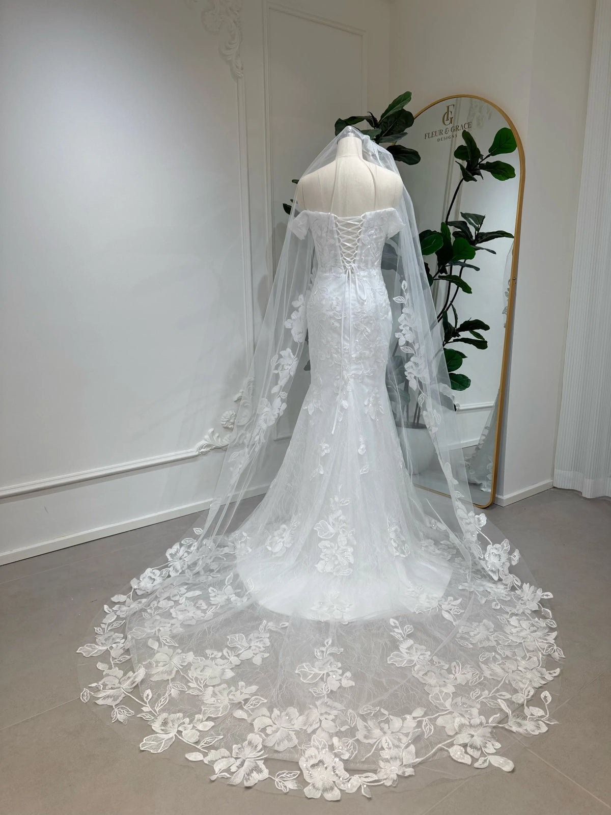 Off-the-shoulder mermaid wedding dress with lace, corset back, and long floral veil on a mannequin.