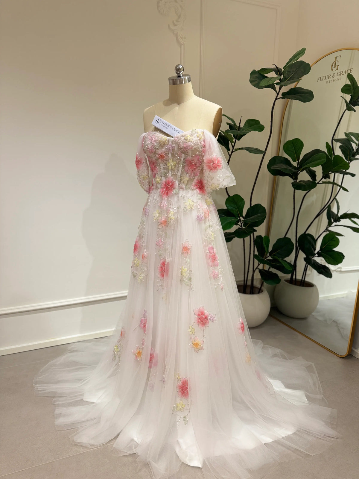 Pink tulle wedding gown with 3D floral appliqués, off-shoulder sleeves, and a chapel train on a mannequin.