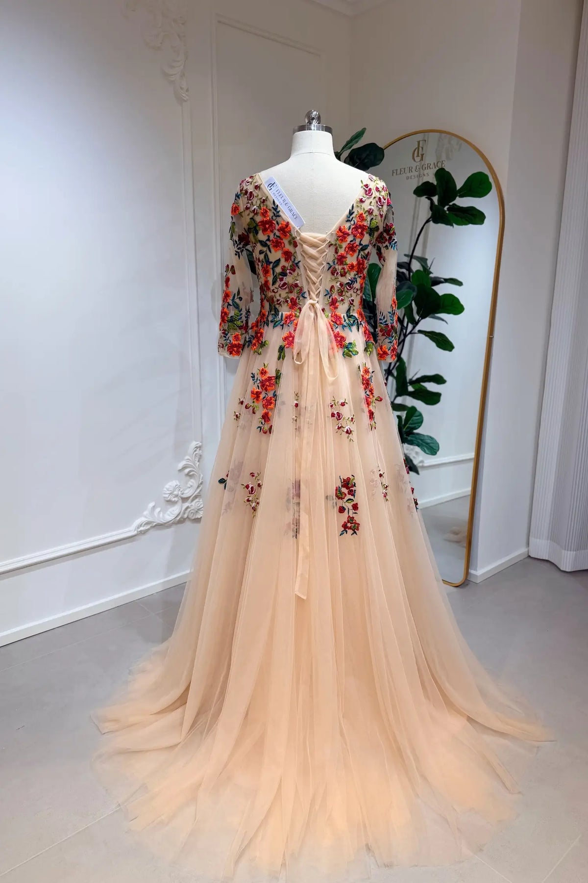 Champagne tulle A-line gown with vibrant orange floral embroidery, 3/4 lace sleeves, and a lace-up back on a mannequin.