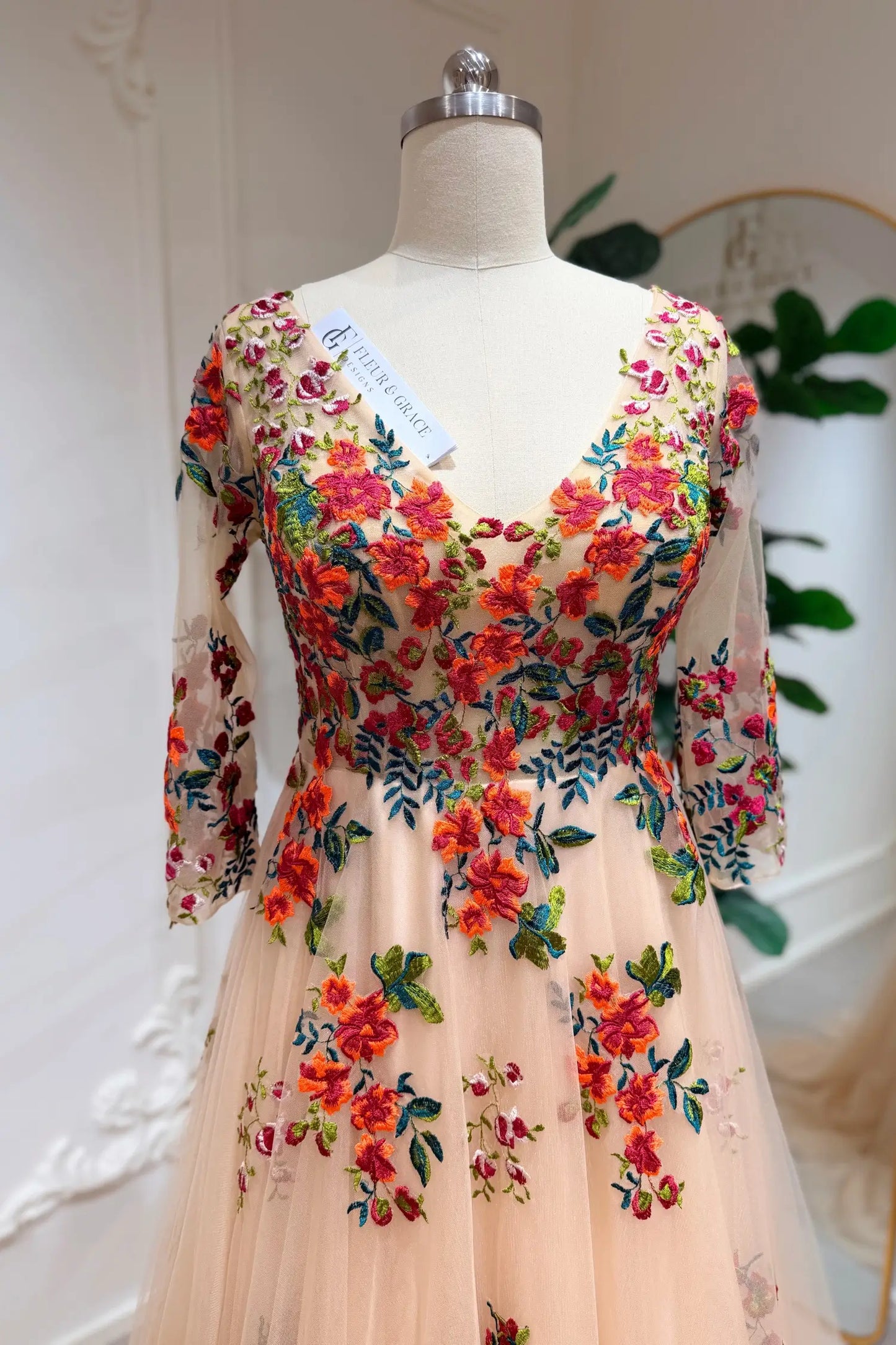 Monica Gown by Fleur&Grace Designs — champagne tulle A-line wedding dress with orange and red floral embroidery, meadow green vines, V-neckline, 3/4 lace sleeves, lace-up back, and pockets for whimsical brides.