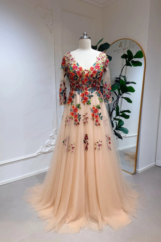Monica Gown by Fleur&Grace Designs — champagne tulle A-line wedding dress with orange and red floral embroidery, meadow green vines, V-neckline, 3/4 lace sleeves, lace-up back, and pockets for whimsical brides.