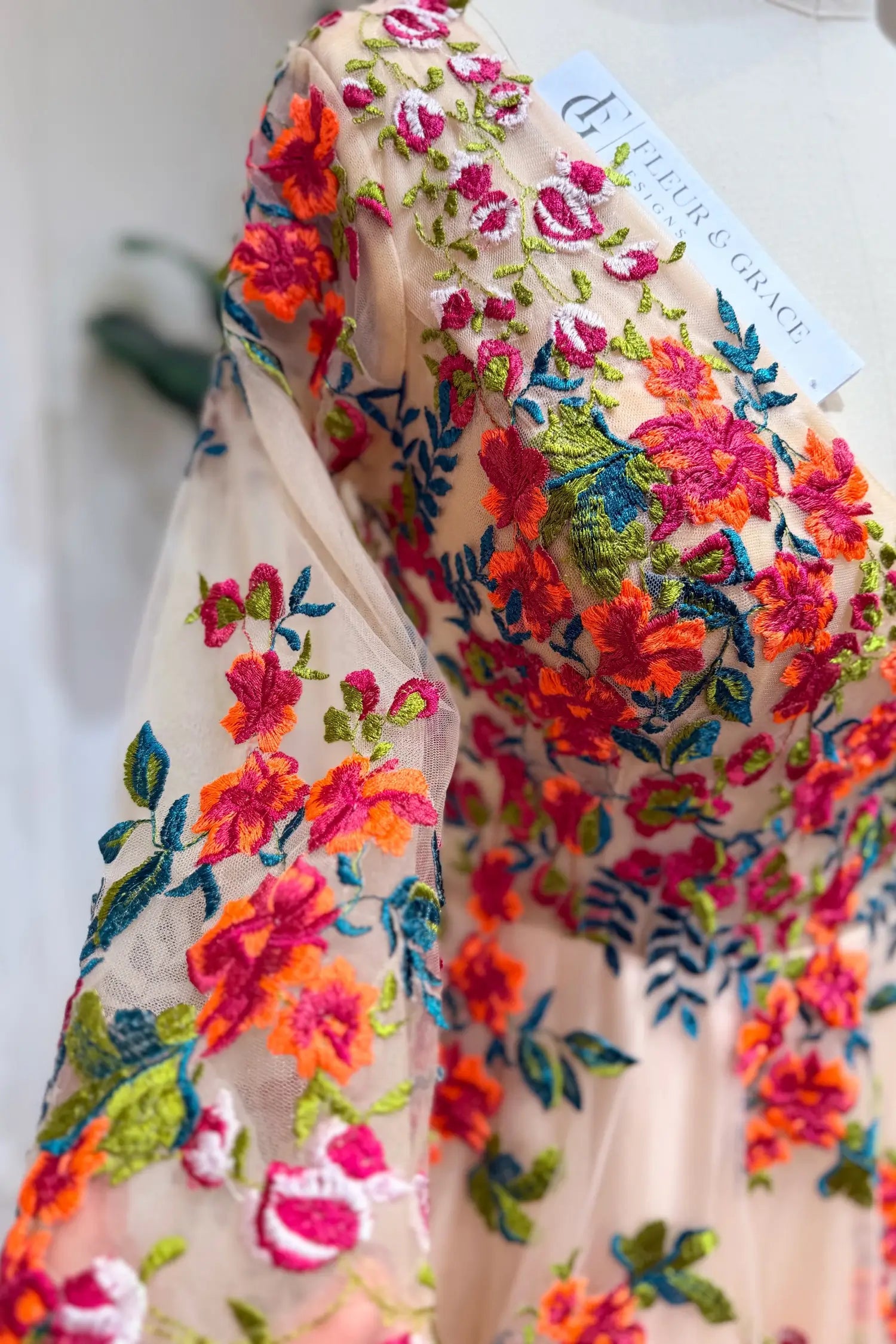 Monica Gown by Fleur&Grace Designs — champagne tulle A-line wedding dress with orange and red floral embroidery, meadow green vines, V-neckline, 3/4 lace sleeves, lace-up back, and pockets for whimsical brides.