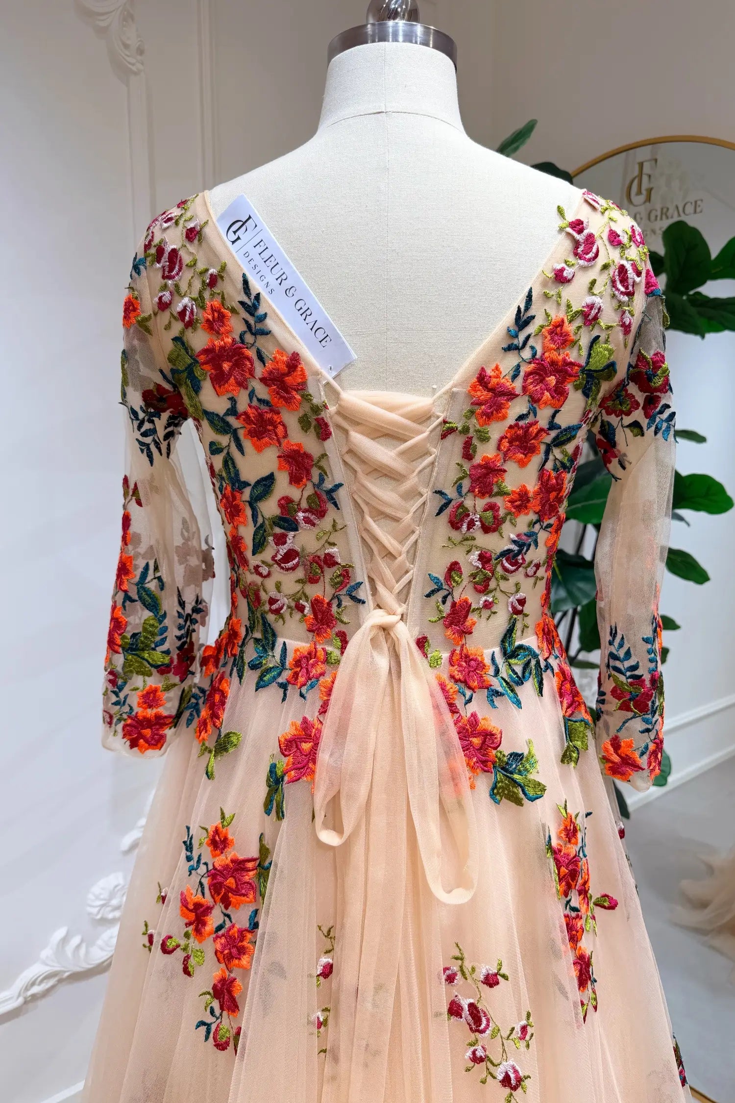 Monica Gown by Fleur&Grace Designs — champagne tulle A-line wedding dress with orange and red floral embroidery, meadow green vines, V-neckline, 3/4 lace sleeves, lace-up back, and pockets for whimsical brides.