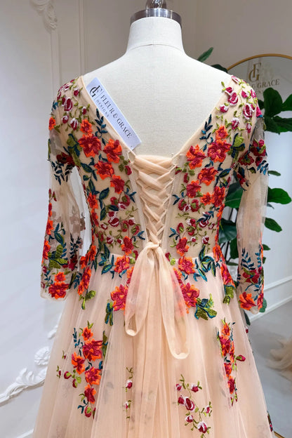 Monica Gown by Fleur&Grace Designs — champagne tulle A-line wedding dress with orange and red floral embroidery, meadow green vines, V-neckline, 3/4 lace sleeves, lace-up back, and pockets for whimsical brides.