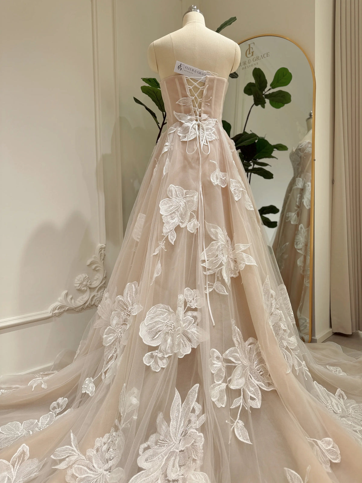 Nude tulle wedding dress with ivory floral appliqués, basque waistline, and lace-up back on a mannequin.