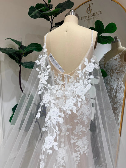 Nola mermaid wedding dress with floral lace appliqué, nude illusion lining, and a dramatic detachable tulle cape.
