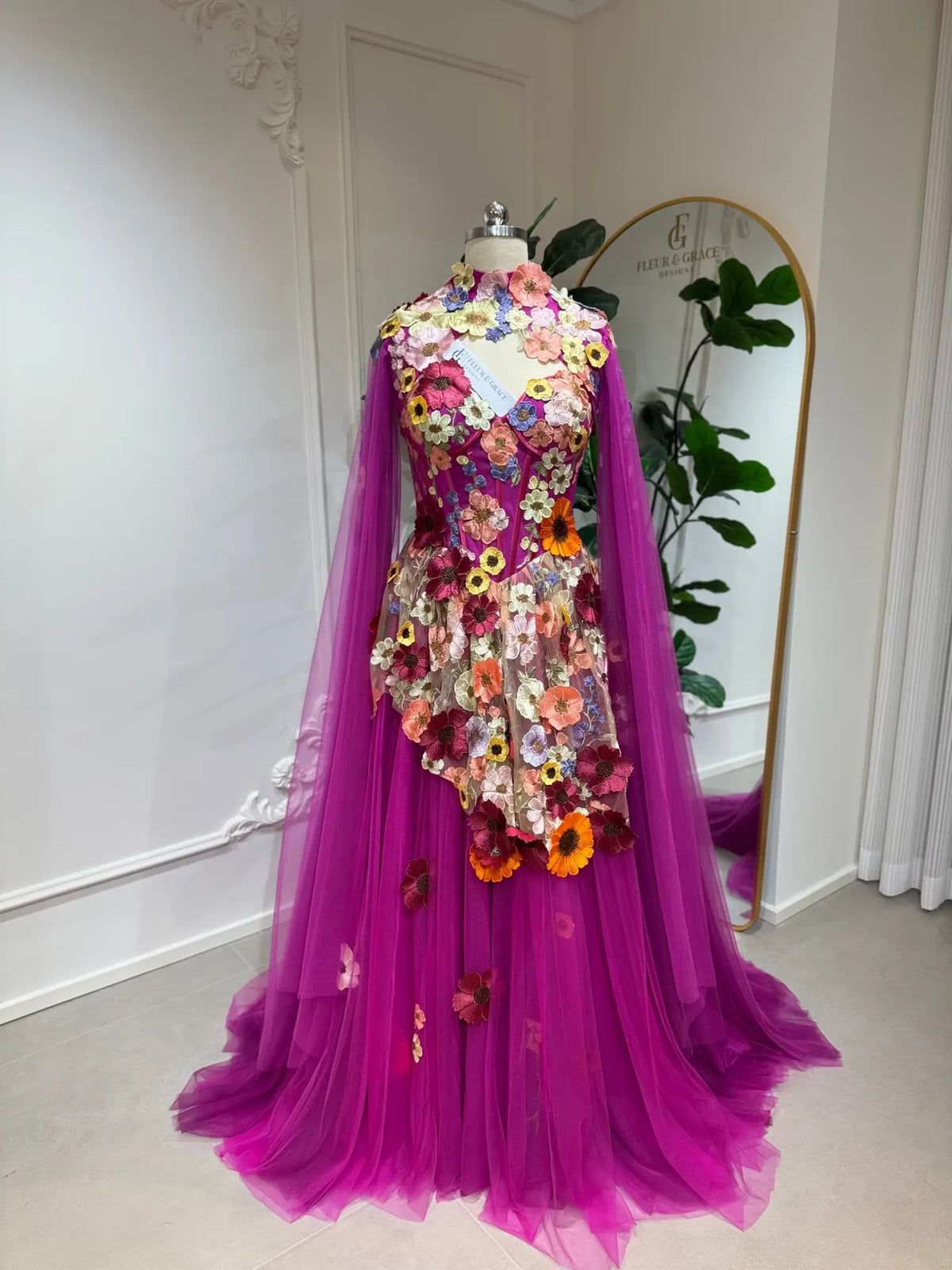 Magenta corset gown with 3D floral appliqués, tulle skirt, and detachable cape sleeves on a mannequin.