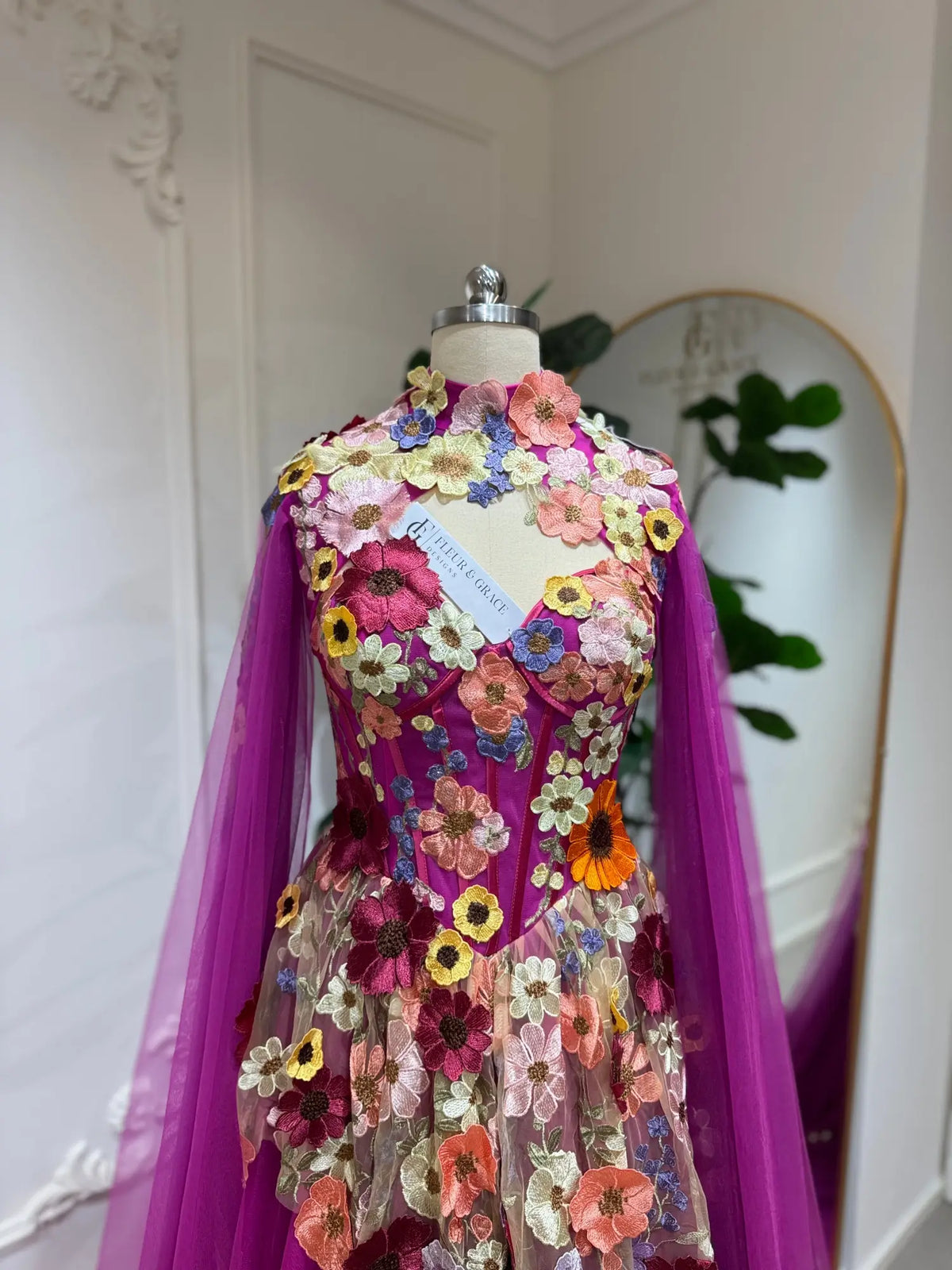Magenta corset gown with 3D floral appliqués, illusion neckline, and detachable sheer cape sleeves on a mannequin.