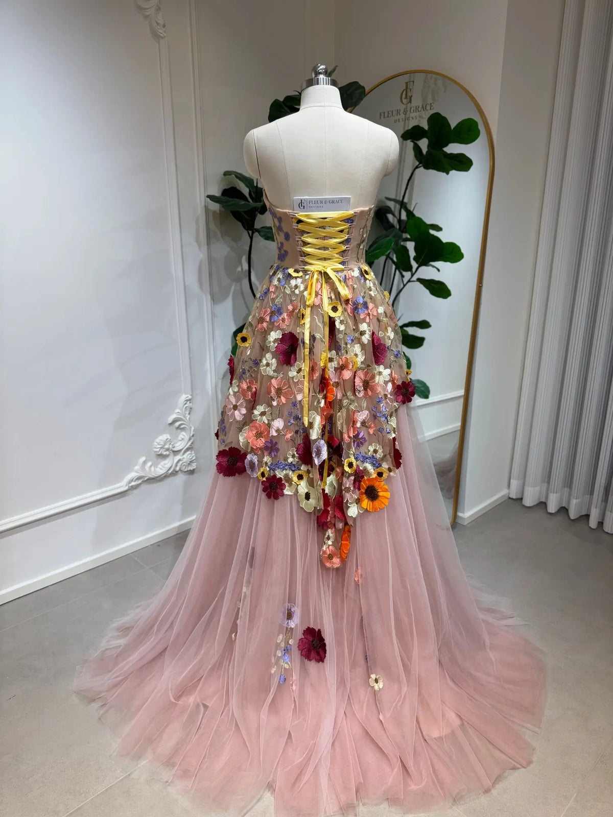 Blush pink tulle ball gown with 3D floral appliqués, corset bodice, and yellow lace-up back detail.
