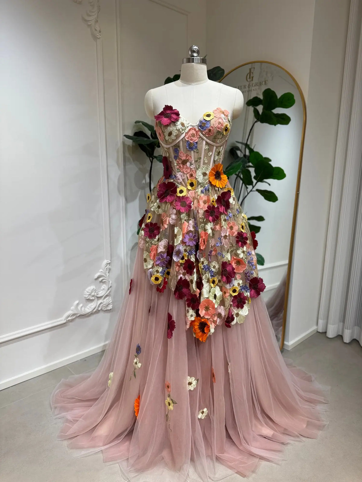 Blush pink tulle ball gown featuring a boned corset bodice and vibrant 3D floral appliqués on an A-line skirt.