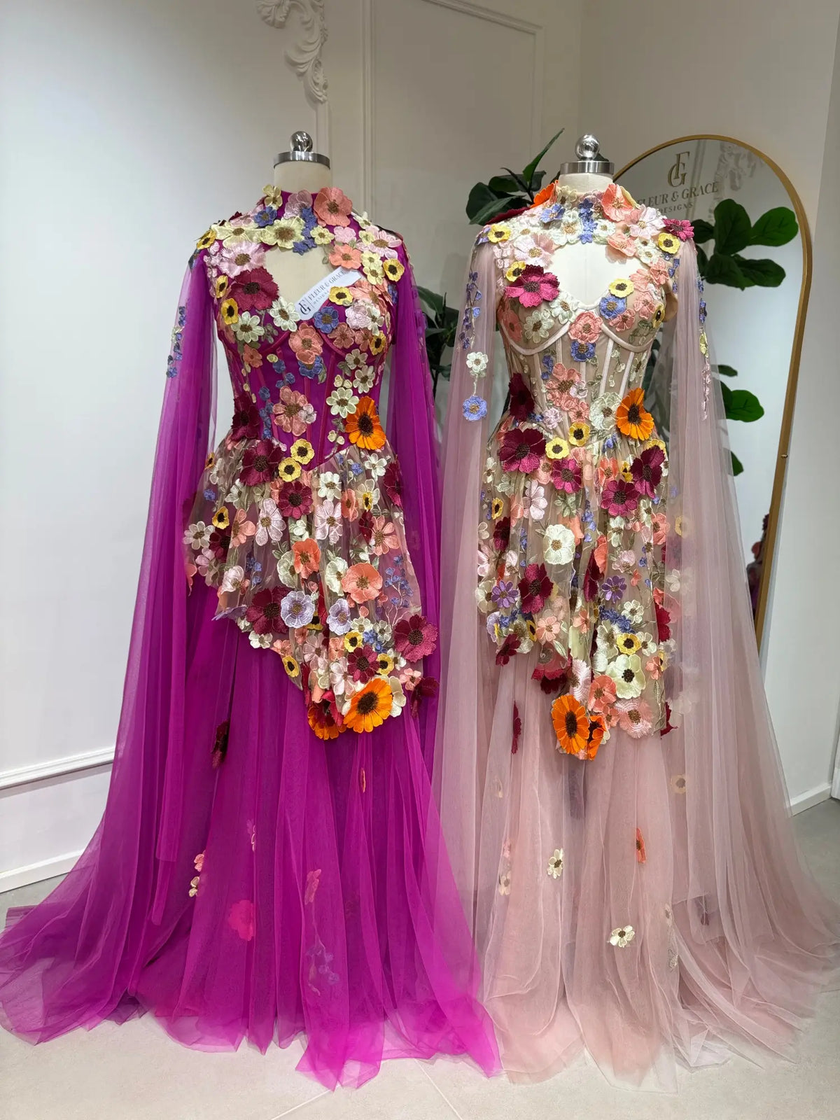 Two Odette floral corset ball gowns in pink and magenta with 3D floral appliqués, tulle skirts, and detachable cape sleeves.