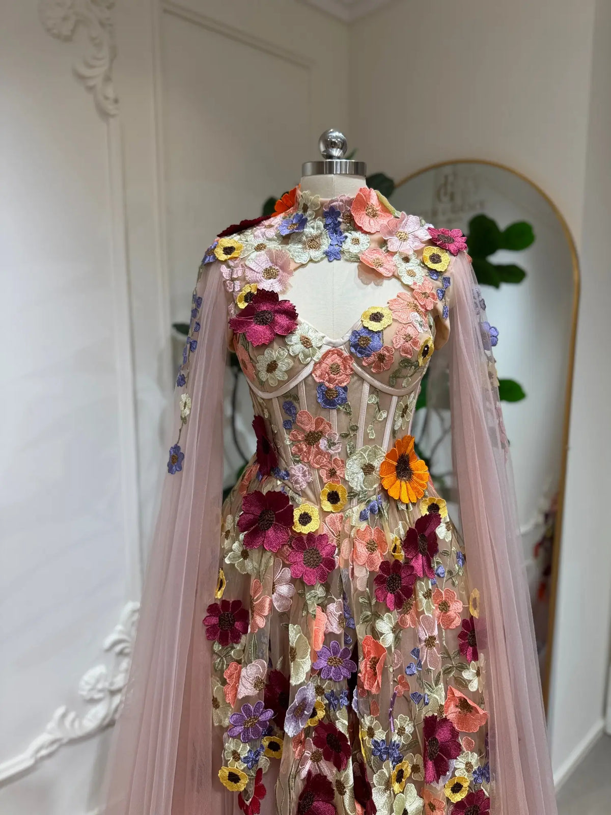 Blush pink corset ball gown with 3D floral appliqués and detachable sheer cape sleeves on a mannequin.