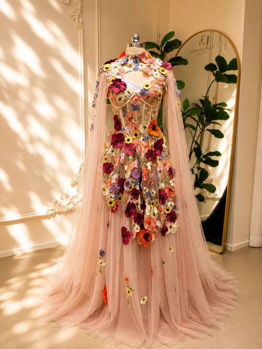 Blush pink tulle ball gown with 3D floral appliqués, a structured corset bodice, and elegant detachable cape sleeves.