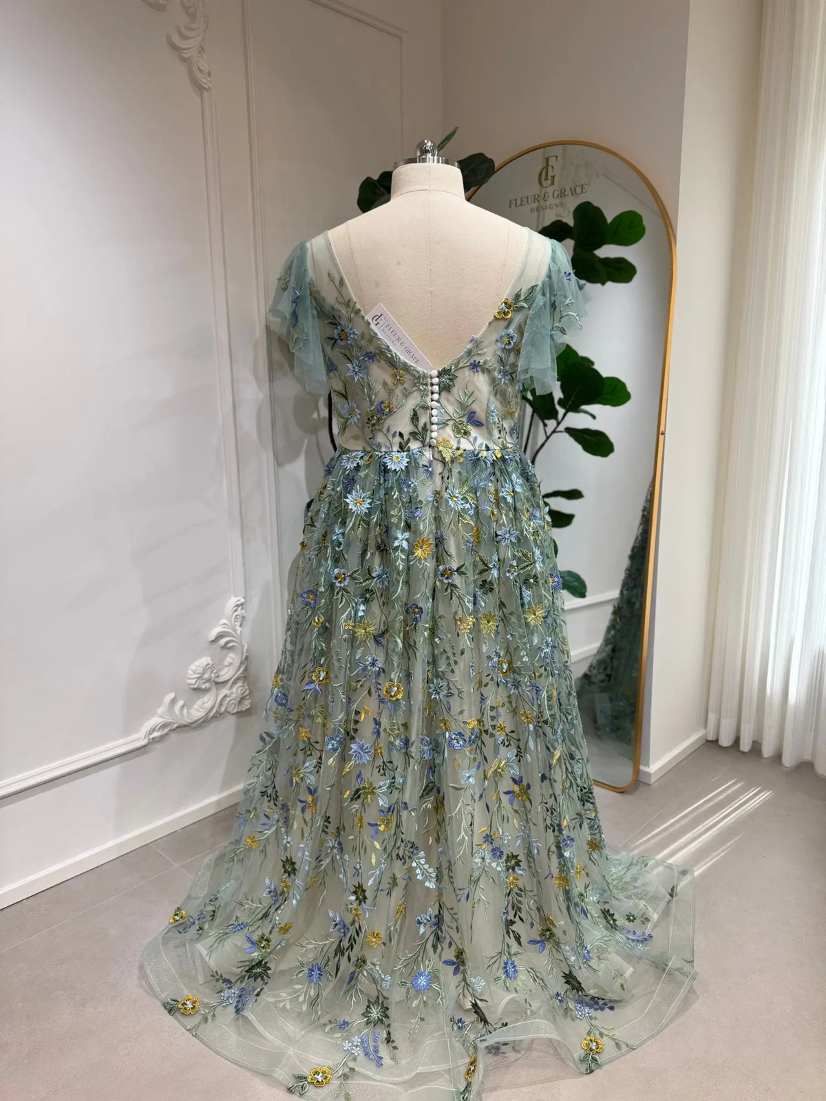 Sage green tulle gown featuring delicate blue and yellow floral embroidery, angel sleeves, and a button-back bodice.