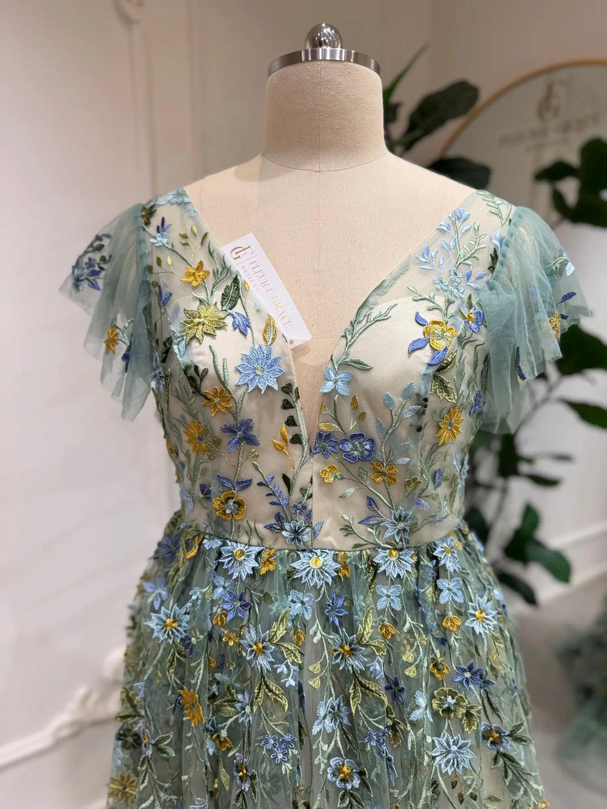 Sage green tulle gown featuring delicate blue and yellow floral embroidery, corset bodice, and whimsical angel sleeves.