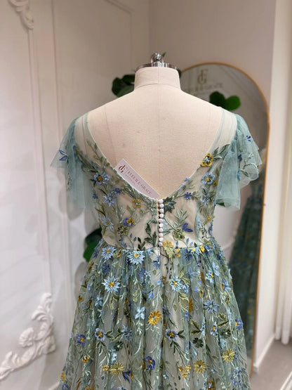 Sage green tulle gown with blue and yellow floral embroidery, sheer angel sleeves, and a button-back corset bodice.