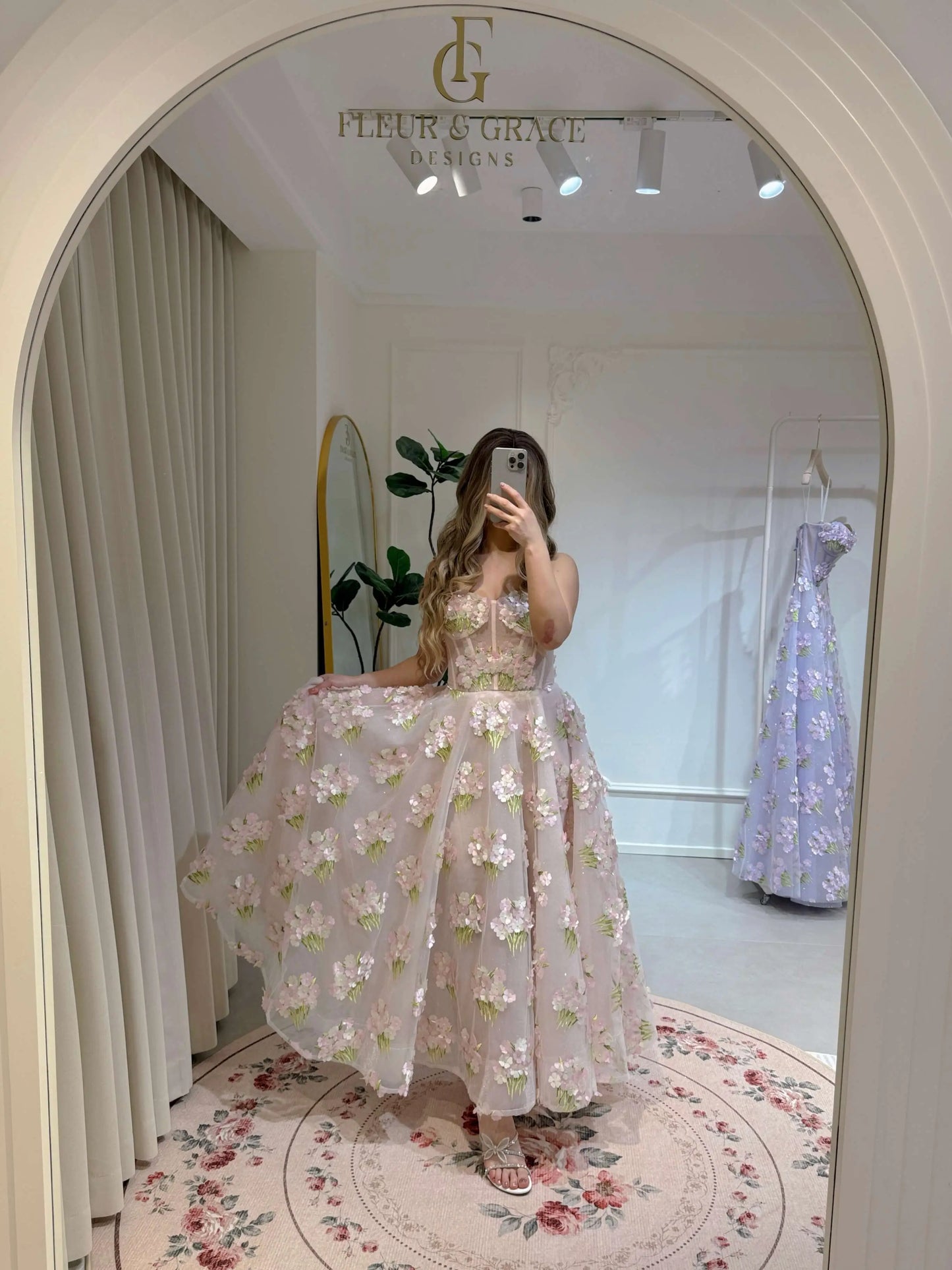 Woman wearing a pink ankle-length gown with 3D hydrangea floral lace and a sparkly bodice, reflected in an arch mirror.