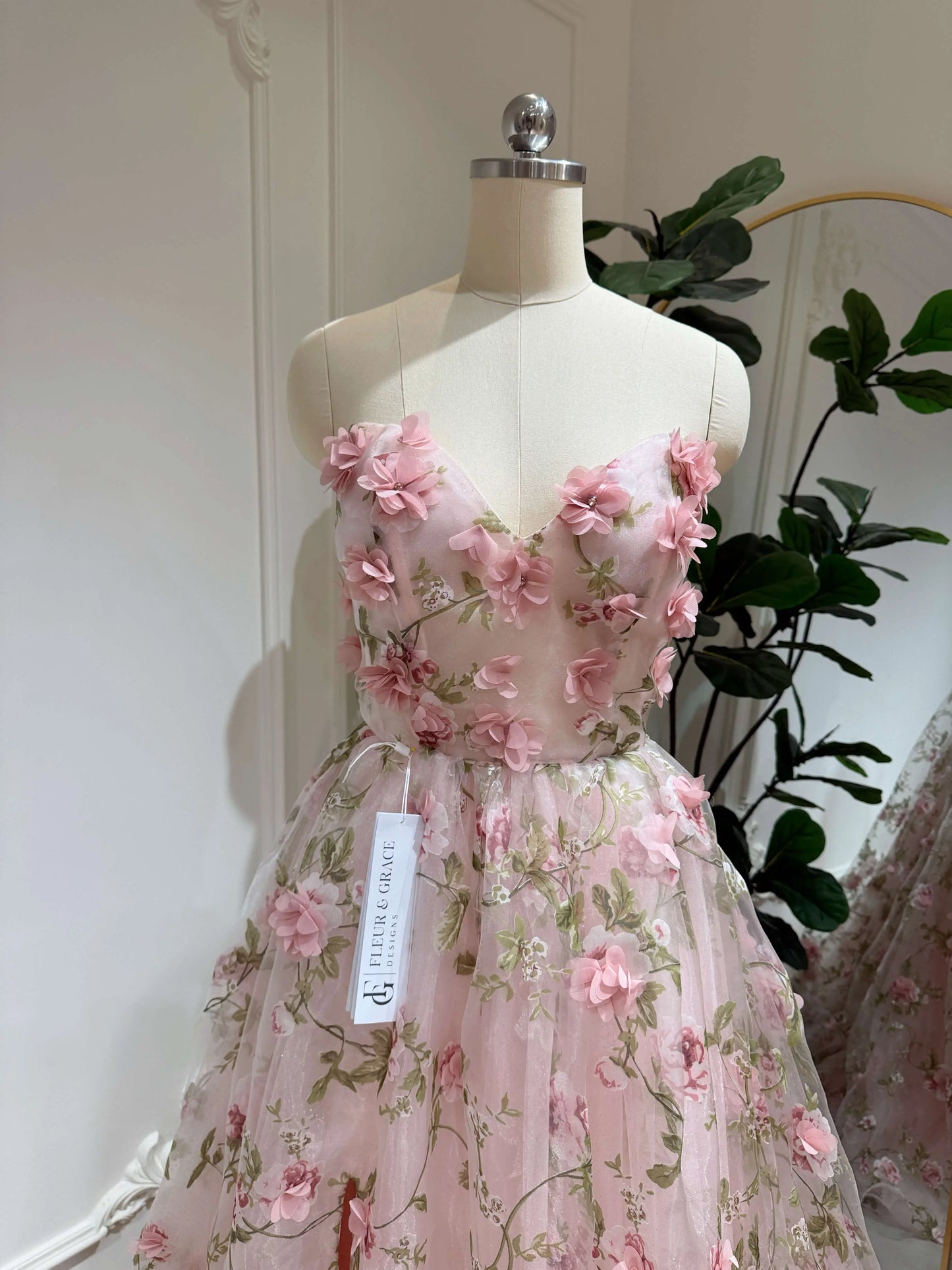 Pink organza prom gown with 3D floral lace, green leaf prints, sweetheart neckline, and sparkly mesh on a mannequin.