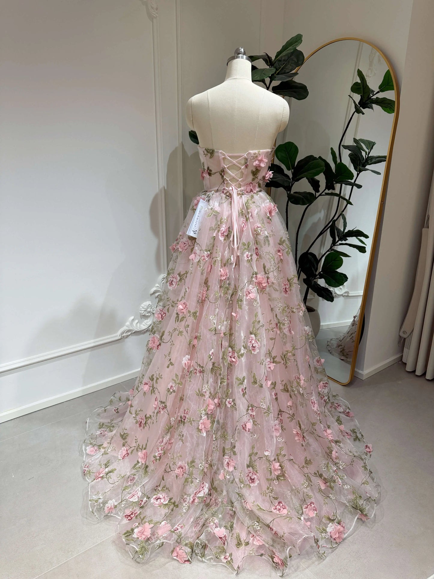 Pink organza gown with 3D floral lace, green leaf prints, and an adjustable lace-up back on a dress form.